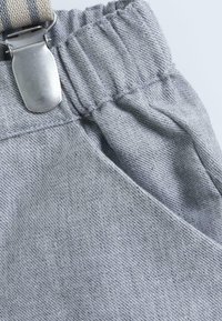 Salopette en tissu gris avec une taille élastique, clip en métal argenté et détails de couture texturés.