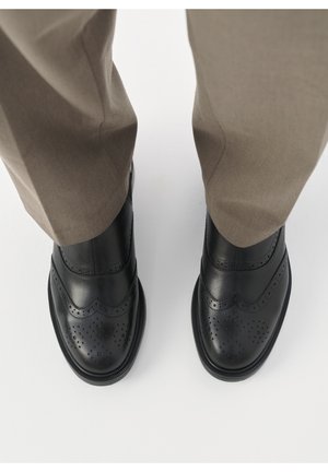 Zwarte leren schoenen met geperforeerde details, een gladde afwerking en contrasterende zolen, gecombineerd met lichtgrijze broeken.