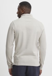 Light beige long-sleeve polo shirt with a stand-up collar, smooth texture, and a side tag; worn, viewed from the back.