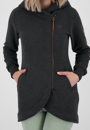 Kobieta ubrana w ciemnoszarą bluzę z kapturem i zamkiem, z małym emblematem w kształcie serca, ręce w kieszeniach, na jasnosiwych spodniach, na jednolitym tle.
