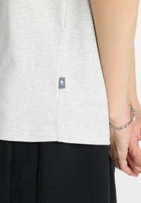 Camiseta de algodón gris claro con dobladillo redondeado. Presenta una pequeña etiqueta azul en el lateral. Llevada con pantalones negros y una pulsera plateada.