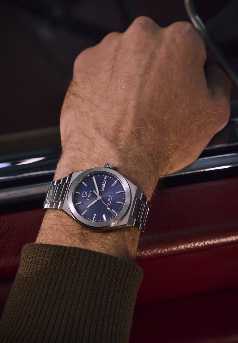 Ceas din oțel inoxidabil argintiu cu un cadran albastru, afișaj pentru dată și curea texturată, purtat pe un încheietură îmbrăcată cu un pulover cafeniu cu dungi.