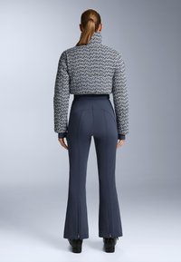 Strukturirana, skraćena jakna u sivo-plavim tonovima s visokim ovratnikom, u paru s mornarskim hlačama s lepršavim nogavicama i patentnim zatvaračima.