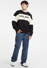 Schwarz- und cremefarbener color-blocked Sweatshirt mit "GUESS JEANS" Logo, kombiniert mit blauen geraden Jeans und weißen Sneakern. Lässiger Schnitt.