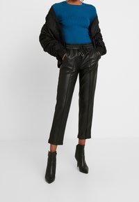Pantalon court en simili cuir noir avec coutures avant, associé à un pull côtelé vert sarcelle et une veste noire matelassée. Des bottines noires complètent le look.