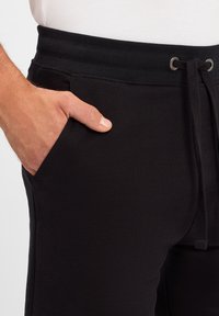 Schwarze Jogginghose mit einem Kordelzugbund, Seitentaschen und einer weichen Textur. Der Stoff erscheint glatt und leicht dehnbar.