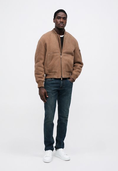 Egy férfi áll, barna cipzáras dzsekit, sötétkék farmert és fehér tornacipőt viselve, egy sima fehér háttér előtt.