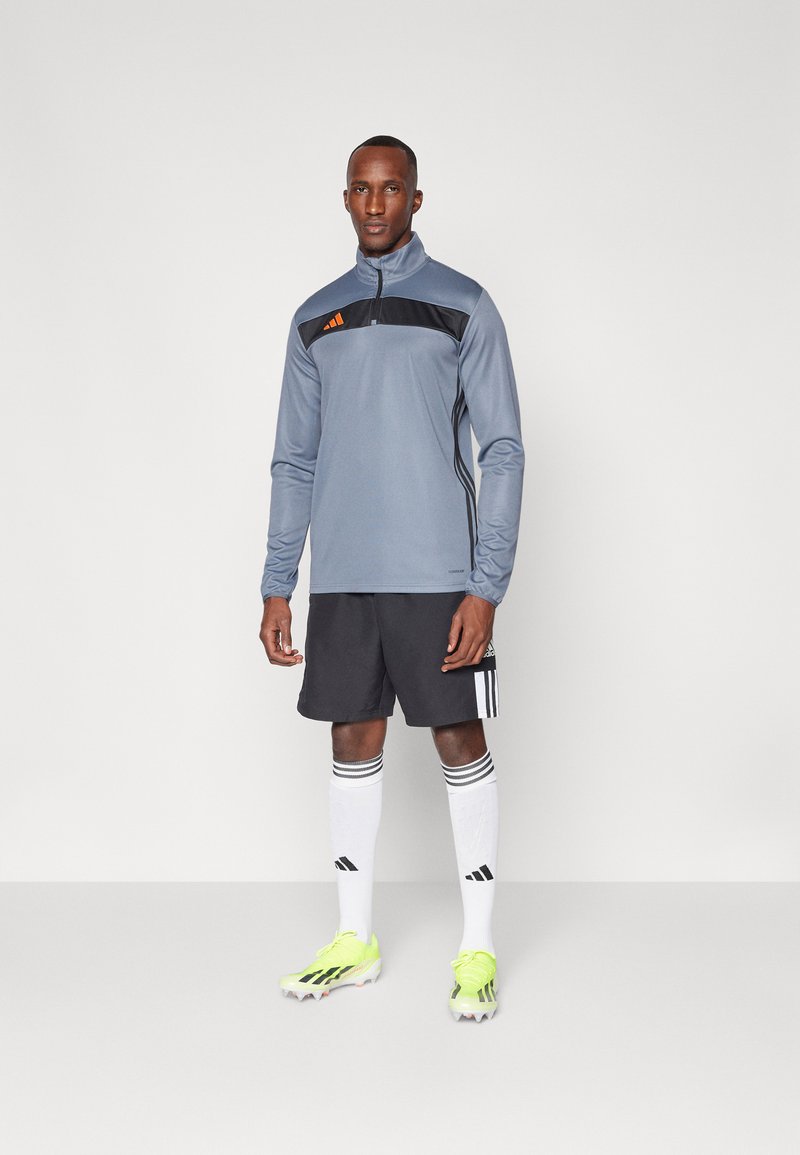 Maglia sportiva grigia a mezza zip con accenti neri, abbinata a pantaloni corti neri, calzini bianchi con strisce nere e scarpe da ginnastica gialle brillanti.