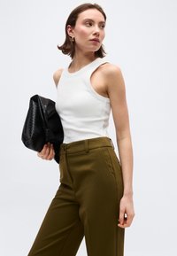 Γυναίκα με λευκό αμάνικο τοπ και χακί παντελόνι, κρατώντας μαύρο υφασμάτινο τσαντάκι, κοιτάζει προς τα πλάγια σε απλό φόντο.