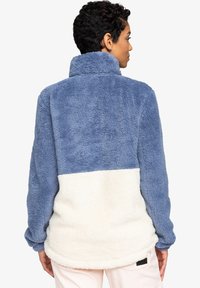 Blå og kremfarget plysj fleece genser med høy krage, med en teksturert overflate og kontrasterende fargeblokkdesign i midjen.