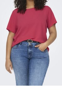 Woman wearing a short-sleeve red top tucked into blue jeans with a black belt, one hand in pocket, against a plain light background.