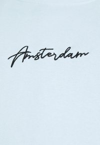 Camiseta de algodón azul claro con texto bordado en negro que dice "Ámsterdam" en una fuente cursiva. Textura suave, corte regular.