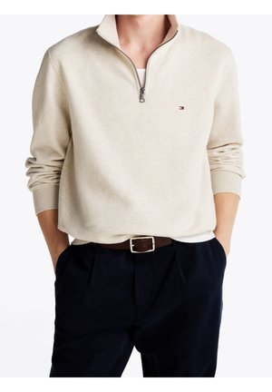 Beige quarter-zip sweatshirt med lille logo på brystet, båret over en hvid undertrøje, parret med marineblå bukser og brun bælte.