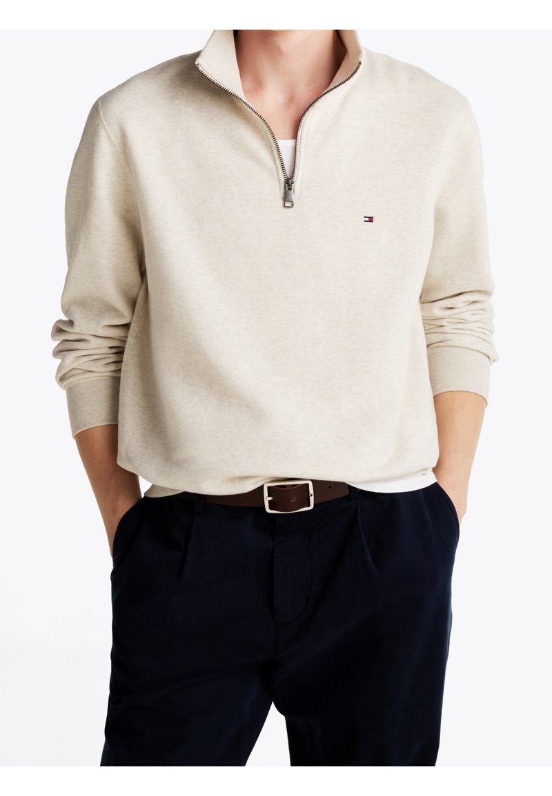 Sweat-shirt beige à col quart-zip avec petit logo sur la poitrine, porté sur un tee-shirt blanc, associé à un pantalon bleu marine et une ceinture marron.
