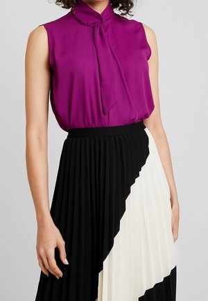 Blusa senza maniche viola con nodo al collo, abbinata a una gonna plissettata bianca e nera con blocchi di colore diagonali. Tessuto liscio.