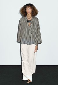 Striped oversized blouse in black and white, paired with loose white trousers. The blouse features a V-neck and wide sleeves.