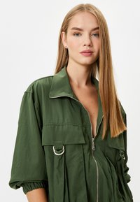 Olijfgroene utilityjack met een oversized pasvorm, voorzien van een rits aan de voorkant, grote klepzakken en accenten van zilverkleurig beslag.