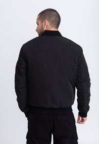 Giacca bomber nera con colletto a costine, polsini ed orlo elasticizzati. Tessuto liscio con cuciture minime, indossata da una persona vista da dietro.