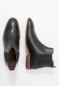 Černé kožené kotníkové boty s elastickými bočními panely, špičatou špičkou a hnědým dřevěným podpatkem. Hladká textura a minimalistické designové detaily.