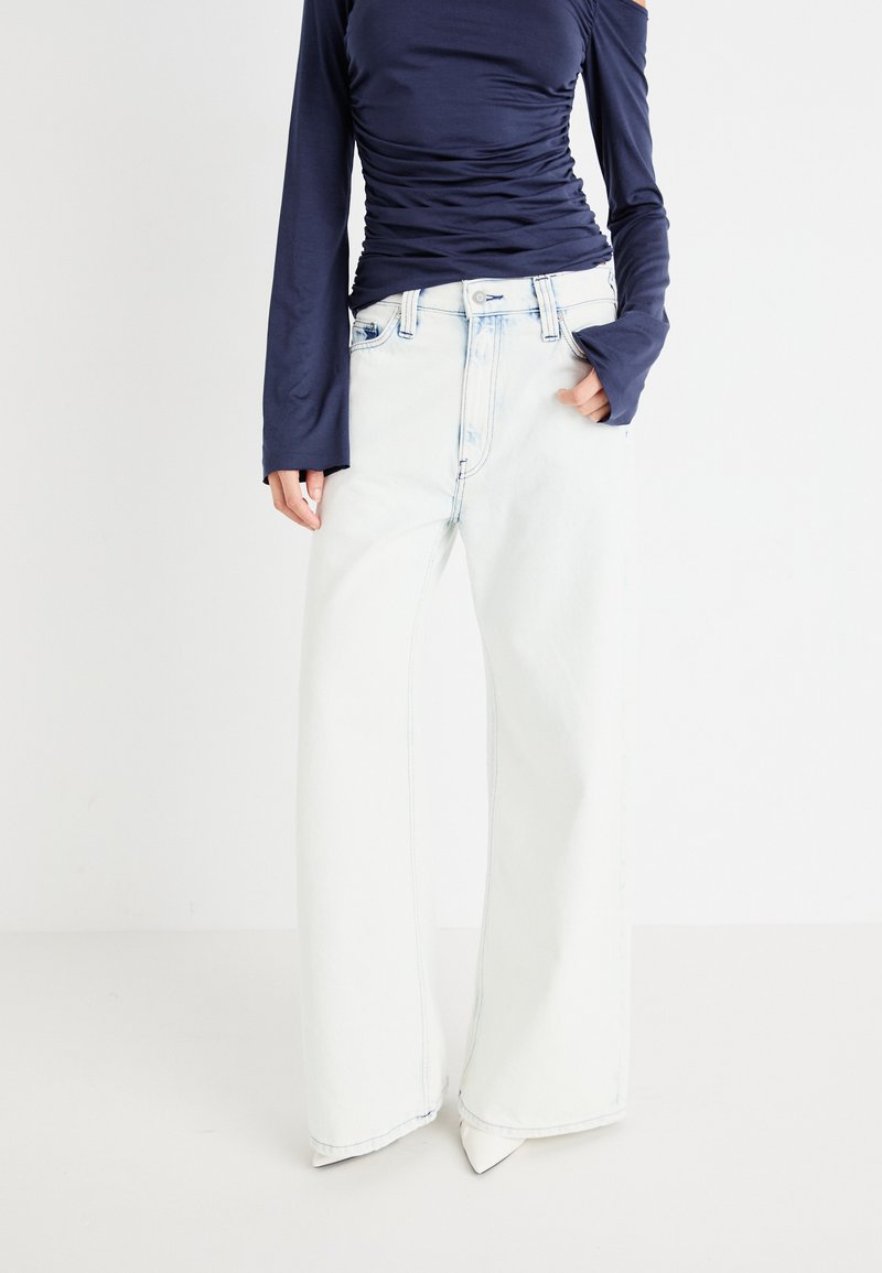 Jeans din denim decolorat, cu picioare largi, de culoare albastră deschisă, cu talie înaltă, finisate cu un cusut contrastant și o textură ușor uzată.
