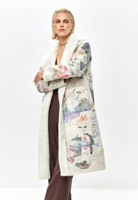 Donna con capelli biondi corti che indossa un lungo cappotto di pelliccia bianca con prints astratti colorati e di animali, abbinato a pantaloni marroni.