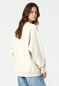 Sweatshirt beige clair surdimensionné en mélange de coton, avec poignets et ourlet côtelés, et épaules basses. Porté avec un jean bleu clair.