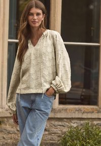 Cremfarbener Pullover mit Kapuze und V-Ausschnitt, im lockeren Schnitt und mit puffierten Ärmeln, kombiniert mit hellblauen Jeans, die mit Nieten verziert sind.