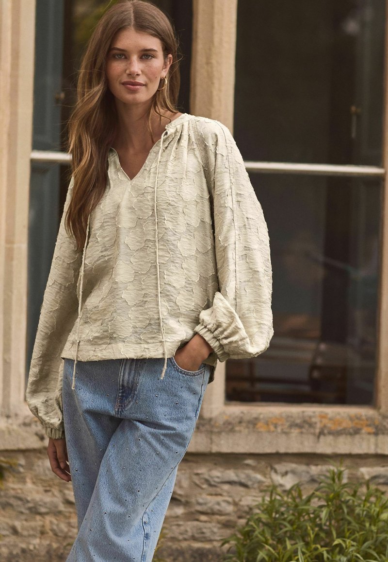 Cremfarbener Pullover mit Kapuze und V-Ausschnitt, im lockeren Schnitt und mit puffierten Ärmeln, kombiniert mit hellblauen Jeans, die mit Nieten verziert sind.