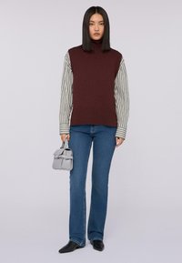 Mujer vistiendo un suéter sin mangas burdeos sobre una camisa a rayas, jeans azules, zapatos de punta negros, sosteniendo un pequeño bolso gris.