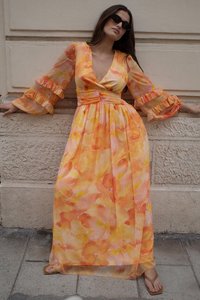 Frau in einem langen, fließenden orange- und gelbblumigen Kleid mit Puffärmeln, Sonnenbrille und Sandalen, die an einer strukturierten Steinmauer lehnt.