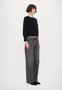 Marineblaue, kurz geschnittene Sweater mit langen Ärmeln, kombiniert mit hochtaillierten, weit geschnittenen grauen Jeans und schwarzen Slippern. Einheitlicher Hintergrund.