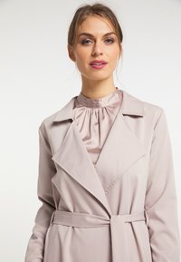 Jasnofioletowy trencz z wyciętym kołnierzem, paskiem w talii i delikatnym połyskiem. Pod spodem bluzka z satyny o wysokiej stójce i zebraniu w dopasowanym odcieniu.
