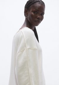 Fehér pulóver környakkal, túlméretezett szabással és nyers szélekkel. Puha anyagból készült, látható varrással és texturált felülettel.