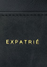 Fekete, textúrázott bőr, amelyen aranyszínű, dombornyomott betűkkel írják, hogy "EXPATRIÉ". Az anyag finom szemcsés mintázattal rendelkezik, ellentétben álló varrással.