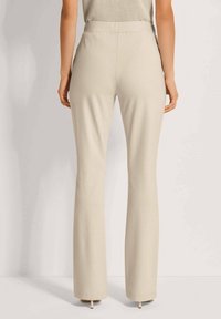 Lichtbeige, rekbare stoffen broek met een gladde textuur, uitlopende pijpen en een aansluitende tailleband. Zicht van achteren, met nadruk op de silhoutte.