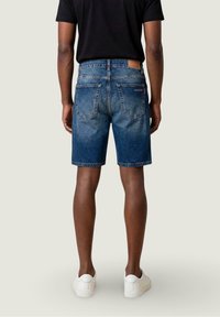 Hombre vistiendo pantalones cortos de mezclilla azul de longitud media, camiseta negra y zapatillas blancas, de pie de espaldas contra un fondo neutro claro.