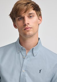 Ljust blå skjorta med knappar gjord av bomull, med en liten marinblå logotyp på vänster bröst, strukturerad krage och synliga framknappar.