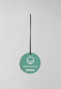 Étiquette ronde verte pour vêtement avec logo écologique blanc et texte « Good Earth Cotton » et « Napapijri Geographic », suspendue par un cordon noir.