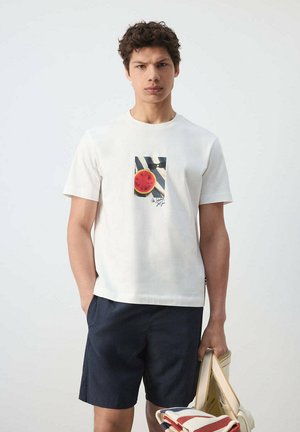 Joven con camiseta blanca con imagen de sandía y pantalones cortos azul marino, sosteniendo una esterilla de playa plegada y sandalias sobre un fondo liso.