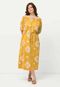 Keltainen olkaimeton mekko, jossa on valkoisia geometrisia kuvioita, kootut kaula-aukot, lyhyet puffihihat ja naru vyötärö. Litteät sandaalit.
