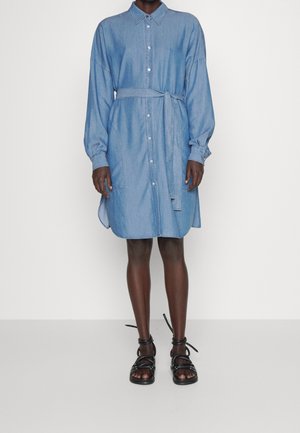 Personne portant une robe chemise en jean bleu clair longueur genou avec manches longues et ceinture à nouer à la taille, associée à des sandales noires à lanières.