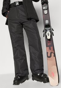 Sorte isolerede skibukser med lige ben, matchende sorte skistøvler og en ski under den ene arm. Bukserne har en tekstureret finish og bælte.