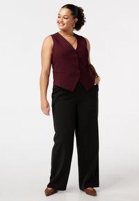 Femme souriante, portant un gilet sans manches bordeaux boutonné, un pantalon large noir, des chaussures pointues à imprimé léopard et des bijoux en or, se tenant avec la main dans la poche.