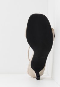 Chaussure à talon haut beige, posée semelle contre un mur et un sol blancs, montrant la semelle noire texturée et l'extrémité étroite du talon.