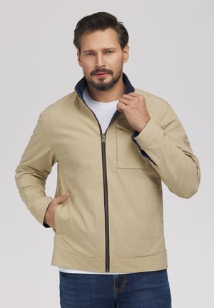 Mężczyzna w beżowej kurtce na suwak, pod którą ma białą koszulę, oraz niebieskich dżinsach, z jedną ręką w kieszeni, a drugą poprawiającą kołnierz, na neutralnym tle.