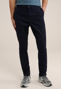 Pantalones de sarga de algodón azul marino con corte recto, bolsillos laterales y cierre de botón. Combinados con zapatillas deportivas grises con suelas negras.