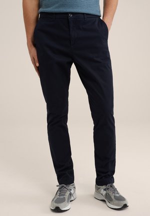 Pantaloni in twill di cotone blu navy con taglio dritto, tasche laterali e chiusura con bottone. Abbinati a scarpe da ginnastica grigie con suole nere.