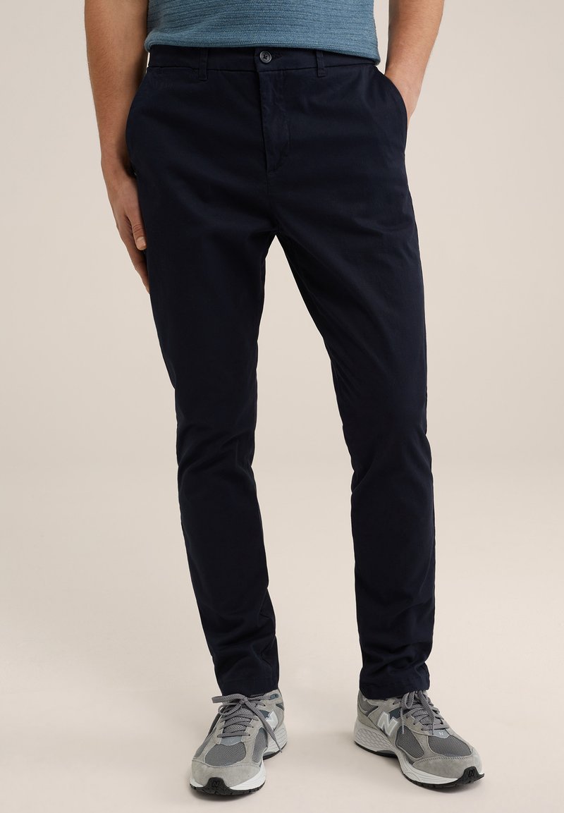 Pantalones de sarga de algodón azul marino con corte recto, bolsillos laterales y cierre de botón. Combinados con zapatillas deportivas grises con suelas negras.