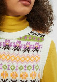 Pull sans manches tricoté avec des motifs floraux multicolores en violet, vert, orange et marron, associé à un col roulé jaune.