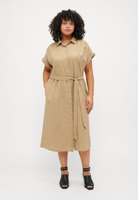 Beiges Leinenhemdkleid mit kurzen umgeschlagenen Ärmeln, Knopfleiste vorne, einem passenden Gürtel in der Taille und Seitentaschen. Tragen mit schwarzen Absatzsandalen.